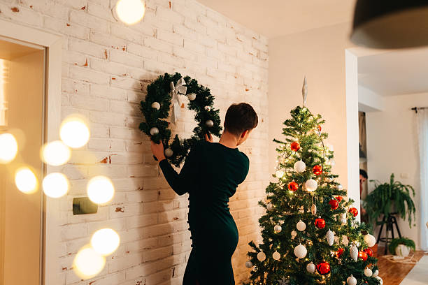 A young woman places a Christmas wreath on a wall, preparing her living space for the holidays. The setting includes a bright tree and glowing festive lights in a cozy atmosphere.