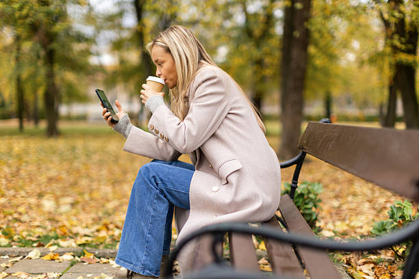istock Woman sitting on a park bench, typing on her phone and drinking coffee 2246160511
