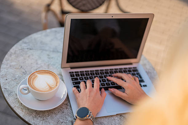Woman hands typing on laptop keyboard with cup of cappuccino on cafe table