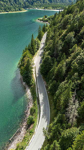 Aerial view of a winding road through a lush forest beside a serene mountain lake, featuring green trees and clear water. The setting provides a picturesque view of nature's beauty, highlighting the harmonious blend of landscape elements.