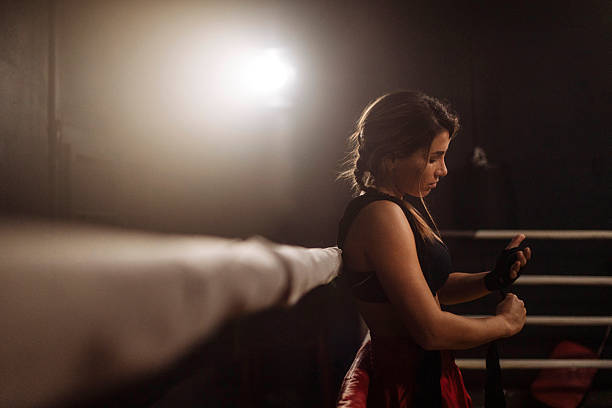 A determined young woman boxer stands in a dimly lit boxing ring, tying her gloves after a training session; The moody light highlights her concentration, strength, and commitment to fitness
