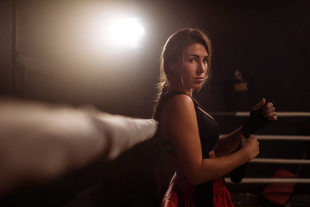 A focused female athlete stands by the boxing ring, adjusting her hand wraps in a moody gym; Soft backlight highlights determination and strength before a training session