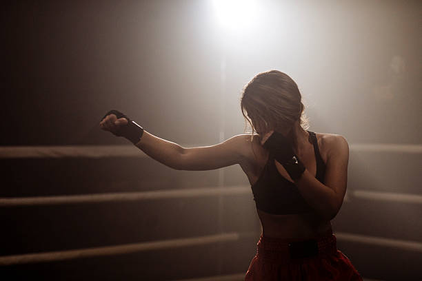A female athlete in a boxing ring throws a punch while wearing gloves and athletic gear; Dramatic lighting emphasizes determination, strength, and discipline during a training moment