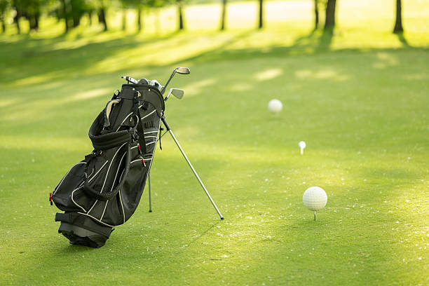 Golf bag standing on a lush green fairway with two teed balls nearby. Sunlight casts gentle shadows on the grass, highlighting the peaceful outdoor setting of the golf course.