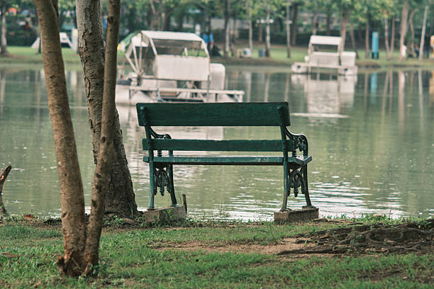 Park bench standing under trees and facing a lake with small boats in the background. Quiet relaxation scene from a Bangkok public park.