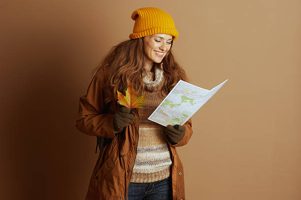 A smiling woman in a yellow knit hat and brown jacket thoughtfully examines a map and an autumn leaf, preparing for an exciting outdoor journey.
