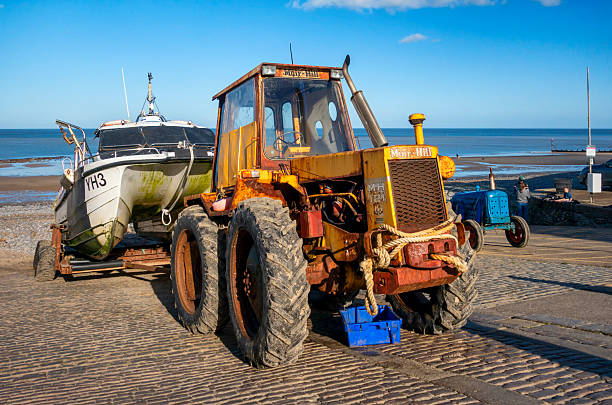 Cromer, Norfolk, United Kingdom: October 18, 2024: A bright yellow but rather rusty beach tractor on a slipway on the beach at Cromer in North Norfolk, Eastern England, on a sunny autumn day. The tractor has pulled a fishing boat up the beach from the surf, with the day’s catch of crabs and lobsters. Cromer is famous for crustaceans.
