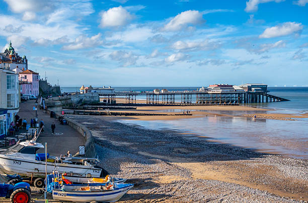 Cromer, Norfolk, United Kingdom: October 18, 2024: An elevated view of the beach at Cromer in North Norfolk, Eastern England, on a sunny day in October. A few people are walking along the beach and fishing boats are lined up above the tide line. The famous Cromer Pier is popular all year round and the shows in its theatre are legendary - along with the crustaceans caught by the fleet of fishing boats. A wind farm can be seen on the horizon.