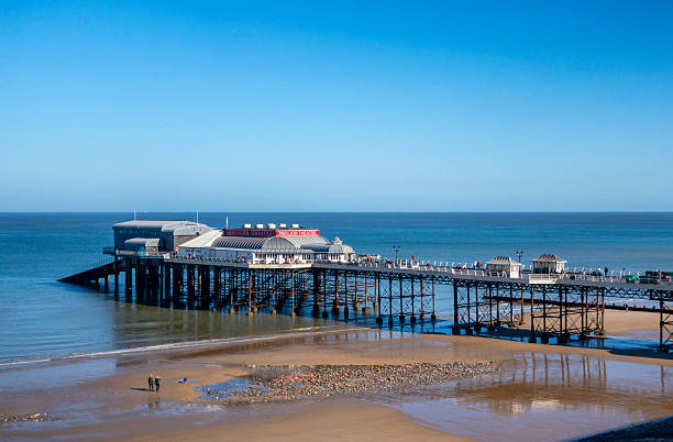 Cromer, Norfolk, United Kingdom: October 18, 2024: A few people strolling amongst the rock pools on the beach at Cromer in North Norfolk, Eastern England, on a sunny day in autumn. Many more tourists and locals are walking on Cromer’s famous pier, with its legendary Pavilion Theatre and lifeboat house. Cromer is famous for much more than crabs!