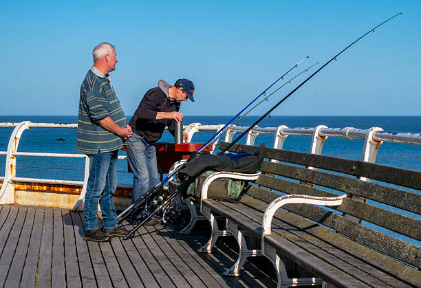 Cromer, Norfolk, United Kingdom: October 18, 2024: Two men with a row of fishing rods at the end of the pier in Cromer, North Norfolk, on a sunny autumn day. Unusually, the North Sea is as blue as the sky.