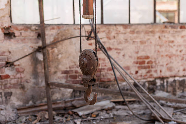 A close-up view of an old metal construction hook hanging inside an unfinished industrial building with exposed brick walls. The image captures the raw atmosphere of renovation and heavy machinery work, symbolizing construction tools, engineering, and industrial progress.