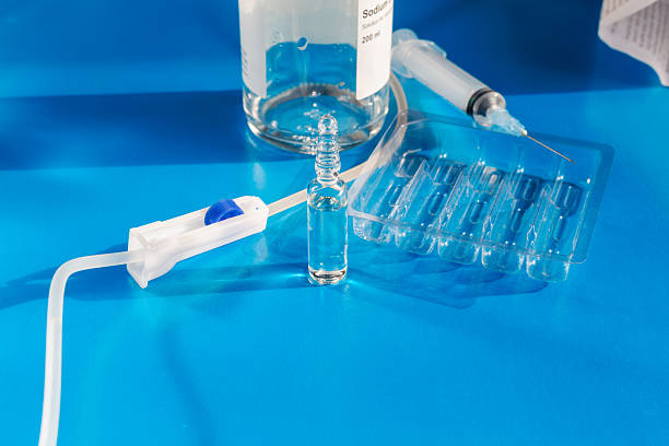 istock Roller clamp on IV drip next to medicine ampoule, syringe, and bottle with saline solution (a mixture of water and sodium chloride) against medication instruction and blue background, close-up. 2246197140