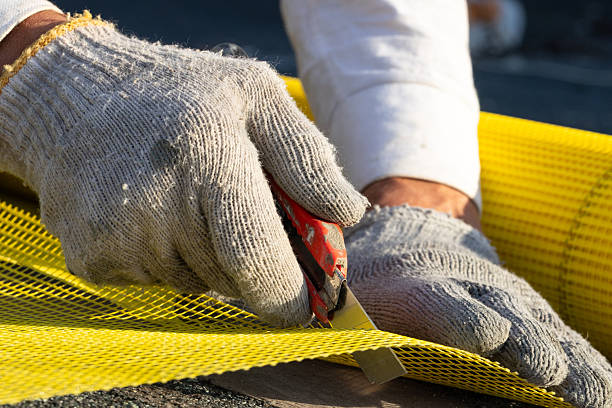 The construction worker holds a sharp utility knife. With his other hand, he holds a piece of reinforcing mesh. The worker cuts the yellow reinforcing mesh in two. The worker prepares the reinforcing mesh for insulating the house's facade.
