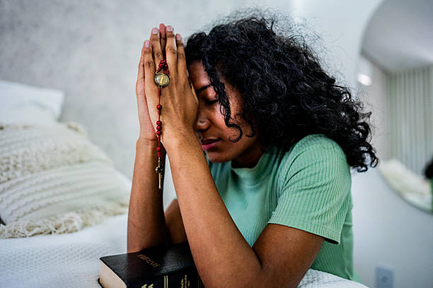 A young woman in casual clothing sits on a bed, engaged in a moment of personal prayer and spiritual reflection, with a bible and holding rosary nearby, suggesting a deeply intimate and contemplative religious experience.