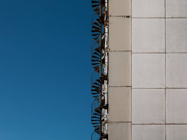 Abstract view of a concrete panel facade and an exterior spiral staircase against a blue sky.