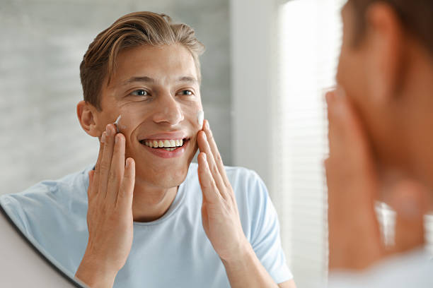 istock Handsome young man applying cream onto his face near mirror at home 2246217921