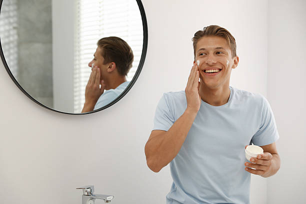 istock Handsome young man applying cream onto his face near mirror in bathroom 2246219111