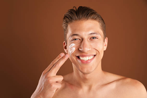 istock Handsome young man applying cream onto his face on brown background, space for text 2246220469