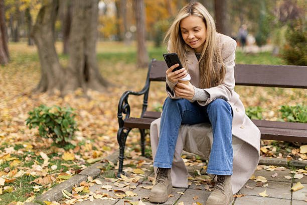 istock Woman sitting on a park bench, typing on her phone and drinking coffee 2246221316