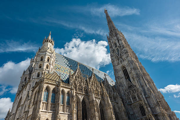St. Stephen's Cathedral in Vienna features intricate Gothic architecture, soaring spires, and a patterned roof, set against a vibrant blue sky with scattered clouds, creating a striking and iconic view of this historic Austrian landmark.