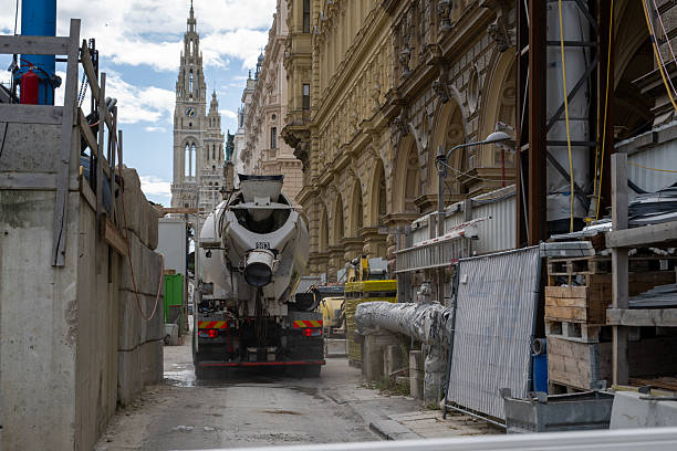 Vienna Votivkirche, a neo-Gothic church with twin spires, under construction with a yellow crane looming overhead. Surrounding buildings, some with scaffolding, frame scene under a cloudy sky