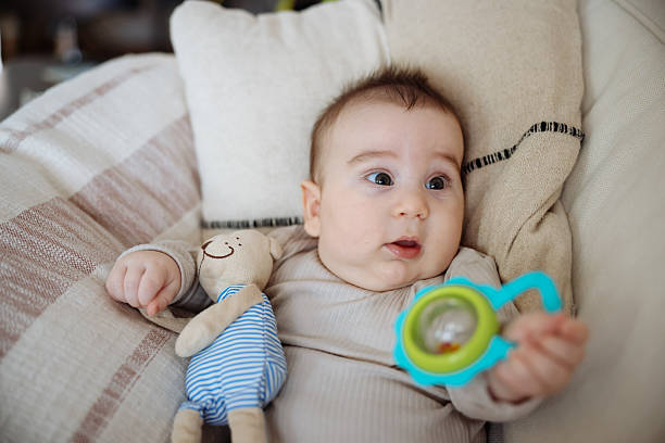 Child plays with toy lying on his stomach. Portrait of cute little baby girl lying with cozy on bed at bedroom, happiness of toddler, newborn 6-11 months with innocence and expression with cheerful, child charm, indoors, one person.