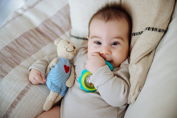 Cute cheerful baby with big light eyes and blond hair sits on bed in playful mood and holds rattle in form of flower, dressed in white crawlers.