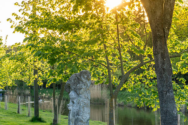 Sunlight shines through fresh green leaves in a serene park, showcasing a stone sculpture beside a peaceful pond during early evening.