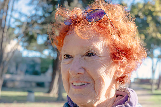 Portrait of a senior woman's face and neck with a background of pine trees, wearing sportswear and orange hair,  smiling.