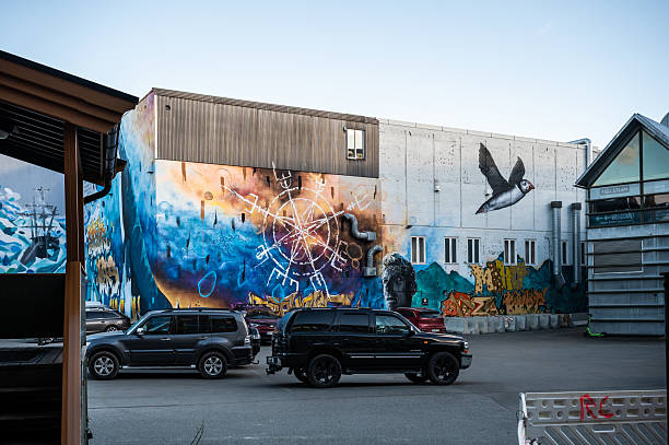 A colorful mural on a building features a bird flying over water, surrounded by celestial symbols, with a statue and maritime elements in the background. Cars are parked nearby, Tromso, Norway