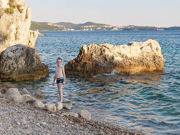 Boy with glasses standing in shallow clear sea by rocks on sunny day. Golden light, turquoise tones, wide copy space. Concept of summer vacation, seaside calm, travel lifestyle.