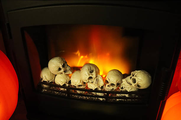 Stock photo shows a spooky Halloween setup with Halloween decorations arranged on a stylish fireplace, complete with a glowing electric fire. Creepy skulls and other eerie decorations enhance the haunted atmosphere