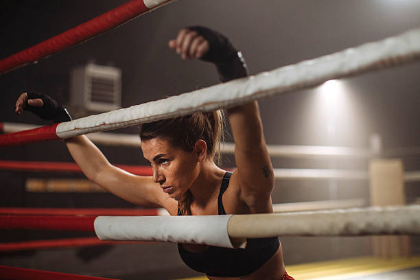 A focused female boxer leans on the ropes of a boxing ring, catching her breath during training; Sweat and determination highlight her athleticism and commitment to fitness