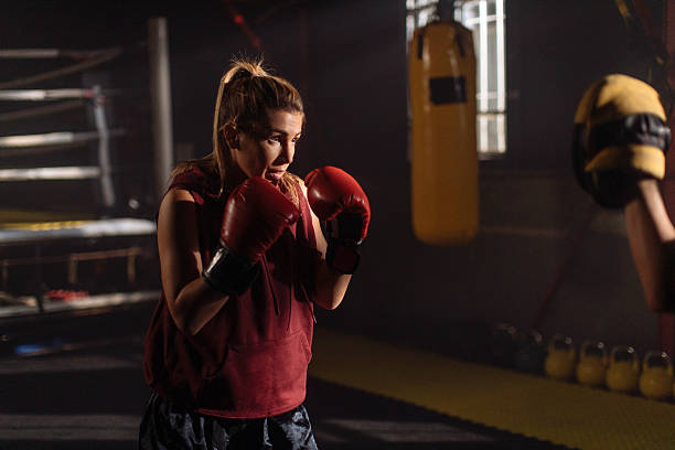 istock Female Boxer Training In Dim Gym With Red Gloves Facing Heavy Bag During Session 2246255155