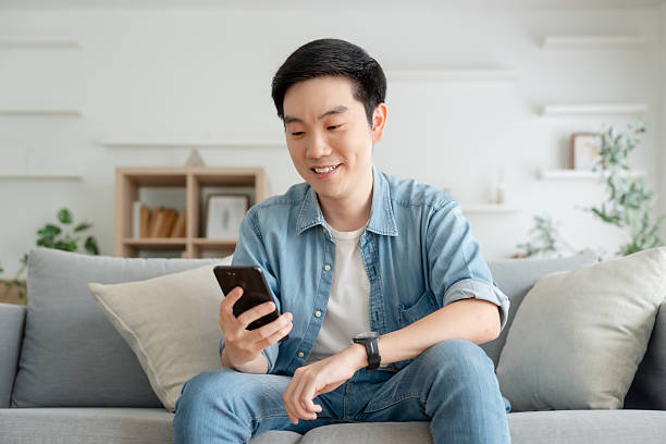 istock Smiling handsome young Asian man sitting on cozy couch, using funny mobile apps in living room. man at home, doing online shopping, messaging friends, posting on social media 2246275767