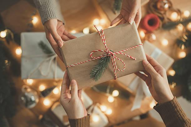 Hands exchanging Christmas gift box wrapped in brown paper with pine decoration and red white string, festive holiday season celebration with warm lights, friendship and happiness in home, New year