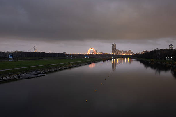 A dramatic twilight view of the Moscow skyline and the illuminated Zhivopisny Bridge, with calm reflections in the Krylatskoye rowing canal under a heavy, overcast sky.