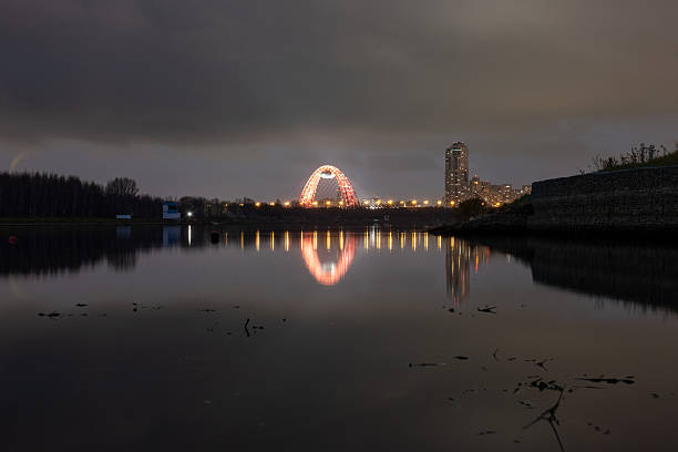 Stunning night view of the iconic red Zhivopisny Bridge in Moscow. Its vibrant lights create a beautiful reflection in the calm water of the rowing canal under a dramatic, cloudy sky.