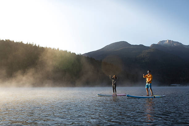 Couple paddle stand up paddleboards on tranquil mountain lake at sunrise, mist rises from Lost Lake, forest and mountains, Whistler, B.C.