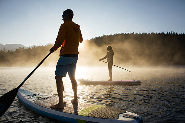Couple paddle stand up paddleboards on tranquil mountain lake at sunrise, mist rises from Lost Lake, forest and mountains, Whistler, B.C.