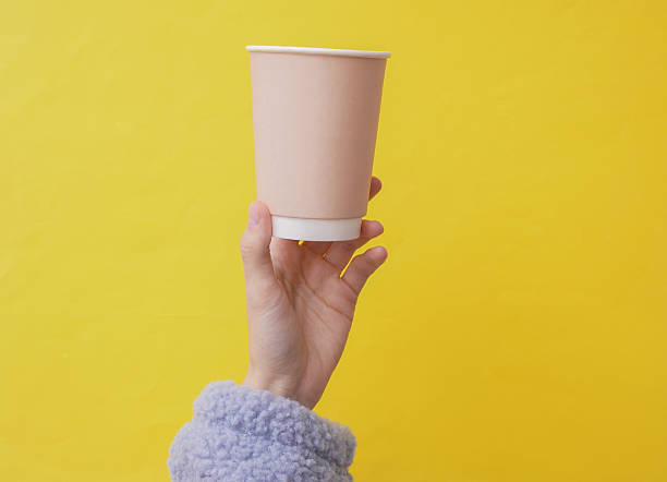 istock Craft eco friendly disposable cardboad cup in a female hand wearing cozy sherpa sweatshirt. Yellow background. Coffee or tea drink 2246297825