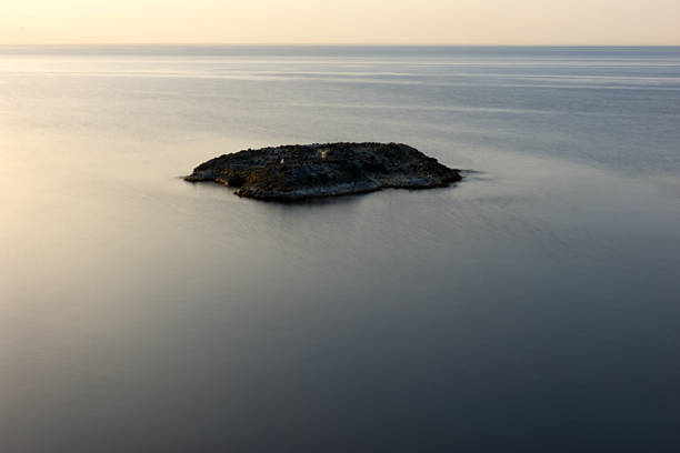 A focused, symmetrical shot of an isolated, dark rock formation resting in the tranquil, glassy water. The smooth texture of the sea surface contrasts with the ruggedness of the rock, all set beneath a diffused, soft light at the horizon. The image conveys a powerful sense of isolation, stillness, and the timeless interaction between water and stone.