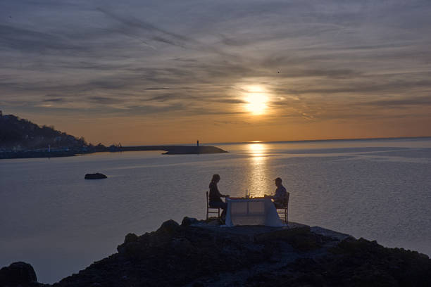 A breathtaking scene of a couple enjoying a private, romantic dinner on a rocky cliff overlooking the calm sea at sunset. The figures and the set table—complete with a white tablecloth and chairs—are silhouetted against the bright, reflective sun path on the water and the dramatic, cloudy sky. This high-impact image captures themes of romance, special occasions, proposal, luxury travel, and unforgettable moments.