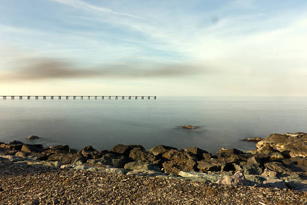 A serene, wide-angle seascape featuring a rough, rocky and gravel shoreline in the foreground, transitioning to the smooth, calm water of the sea. In the mid-ground, a long industrial pier extends into the horizon, adding a geometric line that contrasts with the natural elements. The soft, hazy light near the horizon suggests a late afternoon or early morning, creating a tranquil and expansive mood.