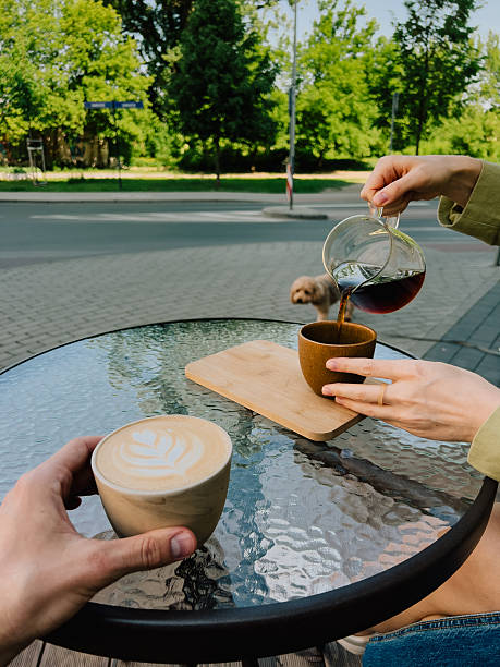 First-person view: couple enjoying coffee at outdoor cafe table, pouring rich coffee into cup while small brown dog sits nearby, basking in warmth of sunny summer day. Pouring coffee into mug