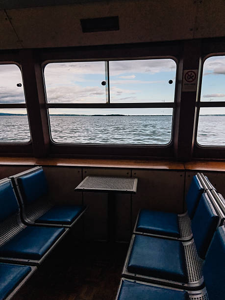 istock Empty passenger seats and small table inside ferry boat navigating on Lake with view on water and cloudy sky through large windows. Vacant ferry seats, small table, large windows revealing Lake waters 2246300768