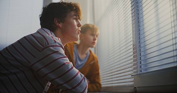 istock Male Students Lean Against Windowsill, Engaged in Calm Conversation While Gazing Through Blinds 2246310726