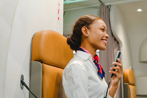 Smiling flight attendant on an airplane jumpseat speaks into a cabin intercom, preparing safety announcements inside a modern aircraft. Inflight cabin airline service business concept
