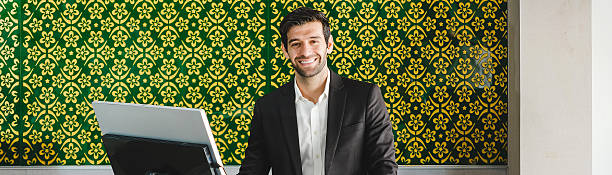 istock Smiling professional in a suit working at a reception desk with a computer, set against a green and gold patterned wall. Airline service business concept 2246311308