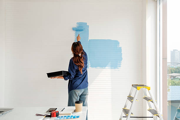 istock Person paints a white interior wall light blue with a roller; paint tray and tools on a table, ladder nearby, city view through the window. 2246311324